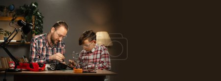 Téléchargez les photos : Père millénaire avec son fils dans des lunettes de protection en utilisant du fer à souder pour réparer la voiture jouet à la maison. Concept de famille et de convivialité. Bannière. Espace de copie - en image libre de droit