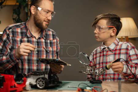 Téléchargez les photos : Père heureux enseignant à son fils pour souder voiture télécommandée à la maison. Espace de copie - en image libre de droit