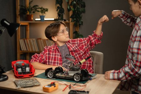 Téléchargez les photos : Des jeunes garçons construisant des robots et des voitures, utilisant du fer à souder pour joindre des copeaux et des fils à la maison. Inventions et créativité pour les enfants. Espace de copie - en image libre de droit