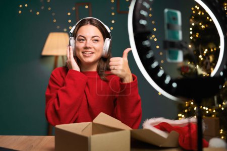 Unboxing video. Caucasian woman in a Santa hat at home shows her subscribers new headphones, listens to music during a live broadcast, raises her thumb up. Influencer advertising concept.