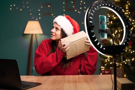 Téléchargez les photos : Blogging, concept de technologie et de personnes - heureuse blogueuse souriante avec anneau lumineux et boîte de déballage smartphone dans le chapeau du Père Noël près de l'arbre de Noël. concept de produits publicitaires avec des blogueurs. - en image libre de droit