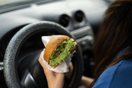 Téléchargez les photos : Femme mignonne conduisant une voiture tout en mangeant un hamburger sur le déjeuner. Attendre et se tenir debout dans un embouteillage. Espace de copie - en image libre de droit