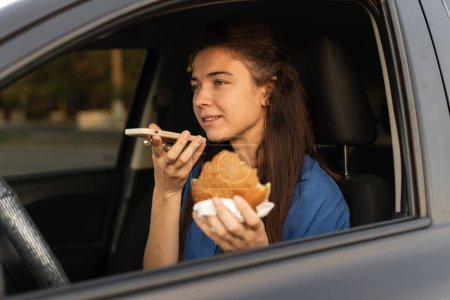 Photo for Focused woman sitting in car when recording voice message on smartphone. Busy lady sending voice message on cellphone and eating burger in car. Copy space - Royalty Free Image