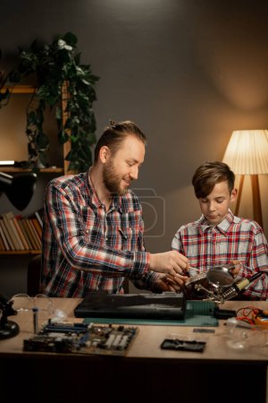 Téléchargez les photos : Jeune père sérieux enseignant à son fils comment réparer un ordinateur portable moderne. Homme et garçon travaillant avec du fer à souder le soir à la maison. Espace de copie - en image libre de droit