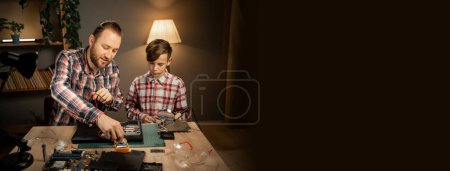 Photo for Father teaching his son to repair a broken laptop and fix the motherboard. Man and child soldering at home in the evening. Banner. Copy space - Royalty Free Image