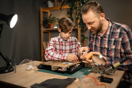 Téléchargez les photos : Père et fils passent du temps ensemble à réparer un ordinateur portable cassé, à l'aide d'un fer à souder et d'autres outils de réparation. Concept de loisirs en famille - en image libre de droit
