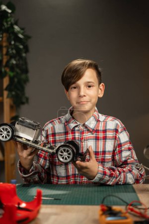 Téléchargez les photos : Portrait d'un garçon réparant une voiture jouet avec des fils à souder regardant la caméra. Soirée à la maison - en image libre de droit