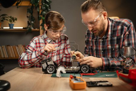 Photo for Father teaching his son for soldering remote controlled car in the evening at home. Man and boy fixing broken toy. Copy space - Royalty Free Image