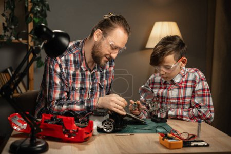 Photo for Father teaching his son for soldering remote controlled car in the evening at home. Man and boy fixing broken toy. Copy space - Royalty Free Image
