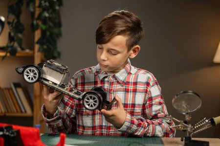 Téléchargez les photos : Portrait d'un garçon réparant une voiture jouet avec des fils à souder regardant la caméra. Soirée à la maison. Espace de copie - en image libre de droit
