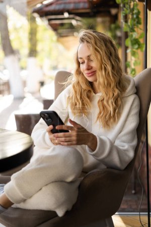 Photo for Beautiful blonde woman in cozy sweater using smartphone and chatting with friends, relaxing at a cafe. Copy space - Royalty Free Image