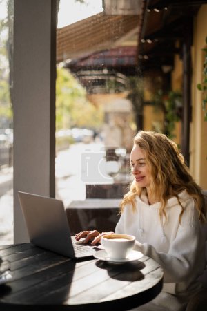 Foto de Mujer rubia escribiendo y viendo en el ordenador portátil en el café sentado en la mesa cerca de la ventana. Concepto de trabajo independiente y remoto - Imagen libre de derechos