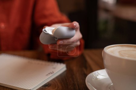 Woman holding headphones case enjoying music through wireless earphones and drinking coffee while sitting in cafe, close-up.