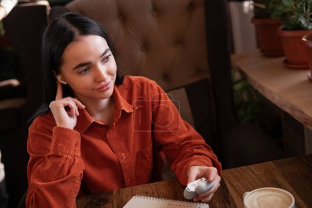 Photo for High angle of girl sitting with earphone in ear holding charging case for wireless headphones while relaxing in cafe. Copy space - Royalty Free Image