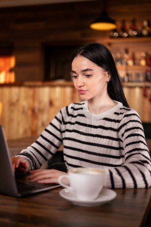 Photo for Portrait of a young woman enjoying coffee during work on laptop, student using computer while sitting in cafe interior during morning breakfast. Copy space - Royalty Free Image