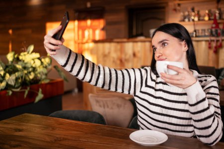 Foto de Preciosa mujer morena sonriente haciendo una selfie mientras toma una taza de café sentado en la cafetería. Copiar espacio - Imagen libre de derechos