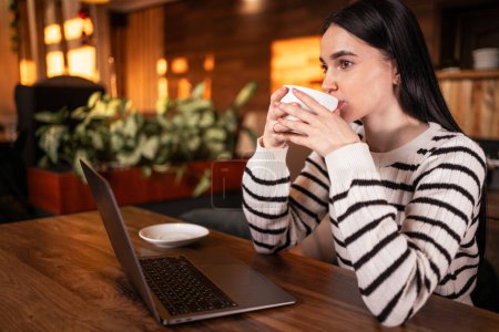 Foto de Joven morena bebiendo café trabajando en línea usando un ordenador portátil sentado en un café mirando hacia otro lado. Copiar espacio - Imagen libre de derechos