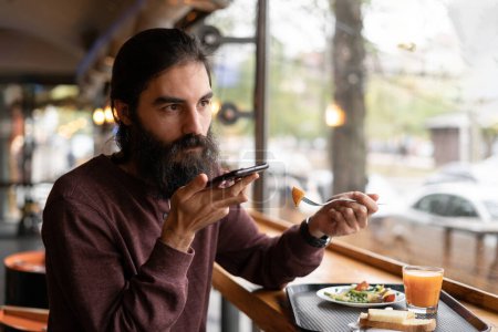 A bearded guy recording a voice message using a smartphone microphone sits in a cafe during lunch break. Copy space