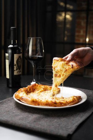 Georgian restaurant. Hand holding a piece of fresh baked cheese-filled georgian flatbread called khachapuri with wine and wineglass on background. Copy space