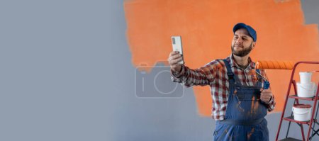 Un pintor masculino haciendo renovaciones en un apartamento nuevo se toma una selfie usando su teléfono inteligente. Banner. Copiar espacio