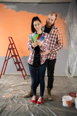 Foto de Pareja joven eligiendo el color para pintar casa de pie cerca de la pared en el apartamento. Reparación, diseño de interiores, construcción, renovación y concepto de hogar - Imagen libre de derechos
