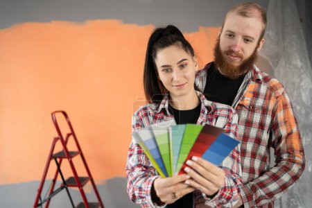 Téléchargez les photos : Jeune couple choisissant la couleur pour peindre la maison debout près du mur dans l'appartement. Réparation, design d'intérieur, bâtiment, rénovation et concept de maison - en image libre de droit