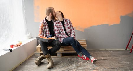 Foto de Alegre renovación de pareja joven en su nuevo apartamento, besándose mientras está sentado en el suelo. Concepto de mudanza y renovación de la casa. Banner. Copiar espacio - Imagen libre de derechos