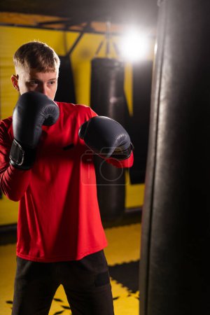 Young boxing male athlete punching boxing bag while exercising in gym during intense workout. Copy space