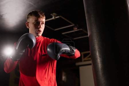 Attractive male punching a bag with boxing gloves. Boxer training in boxing gym. Low angle