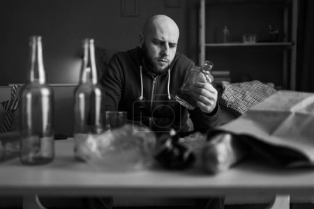 Millennial einsamer depressiver Mann, der zu Hause Whisky trinkt und am Tisch mit Glas sitzt. Alkoholmissbrauch, Alkoholismus Konzept