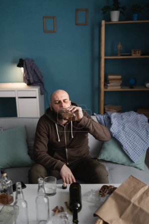 Téléchargez les photos : Seul triste homme déprimé buvant du whisky à la maison assis dans un salon désordonné. Concept de dépendance et d'alcoolisme. Espace de copie. Angle élevé - en image libre de droit