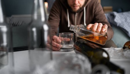 Nahaufnahme eines Alkoholikers, der Whiskey in ein Glas auf einem Tisch gießt, während er im Wohnzimmer zwischen Flaschen und Schmutz sitzt. Alkoholsucht und psychische Probleme