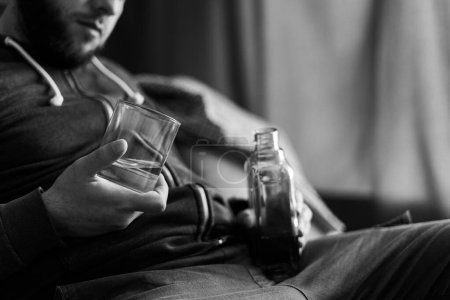 Betrunkener und einsamer Mann mit Whiskeyflasche und Glas auf Sofa sitzend, in Großaufnahme. Alkoholmissbrauch, Alkoholismus Konzept. Schwarz-Weiß-Foto