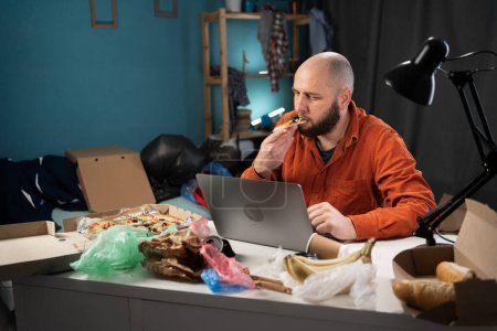 Photo for Bearded man eating pizza and working in messy, cluttered freelancer room with piles of clothes. Copy space - Royalty Free Image
