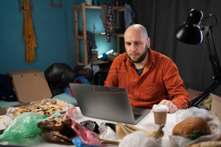 Busy male freelancer using laptop surrounded by tons of garbage and food in his apartment. Copy space