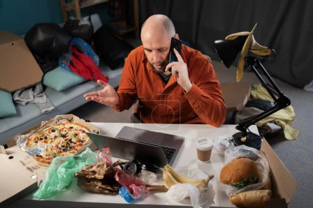 Slatternly bearded man sitting before laptop and holding smartphone among leftovers, food, bags and rubbish on background. Copy space