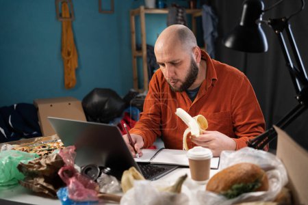 Freelancer working at home in dirty cluttered room, man using laptop computer eating banana at workplace. Copy space