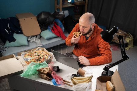 Photo for Bearded freelancer man working at home, eating pizza in Messy, cluttered room with piles garbage. Copy space - Royalty Free Image
