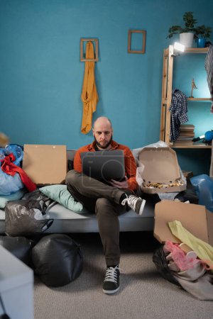 Busy male freelancer using laptop sitting on sofa surrounded by garbage in his apartment. Copy space