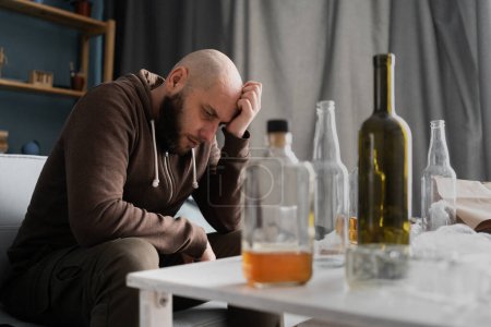 Alcohol addiction abuse and alcoholism concept. Upset millennial man drinker alcoholic sitting at home with empty bottles drinking whiskey alone, sad depressed addicted man having problem.