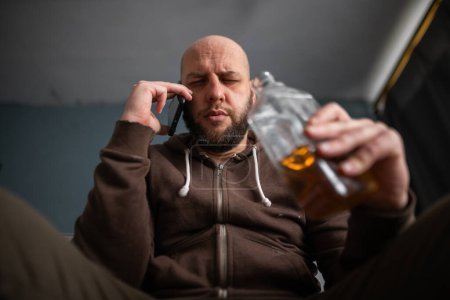 Photo for Drunk alcoholic man drinking whiskey from the bottle and calling on mobile phone depressed at home. alcohol abuse and alcoholism concept. Low angle - Royalty Free Image