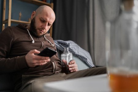 Téléchargez les photos : Dépendance à l'alcool et concept humain. Homme ivre avec smartphone et bouteille de whisky assis sur le canapé à la maison. Espace de copie - en image libre de droit