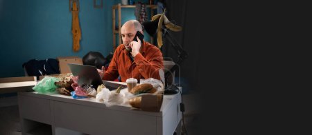 Photo for Man working in Messy, cluttered room. Bearded freelancer making phone call and using laptop at home. Banner. Copy space - Royalty Free Image