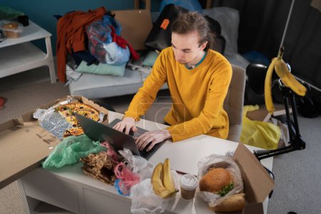 Photo for Hipster student studying and working in a dirty messy room using a laptop among stacks of clothes. Copy space - Royalty Free Image