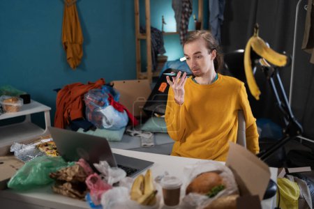 Young man working studying in messy room dictate voice message on smartphone. Copy space