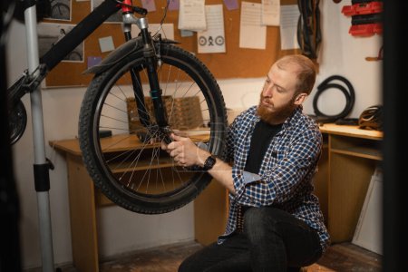 Bearded red-haired man in a checkered shirt repairs a bicycle while squatting in a garage or workshop. Side view