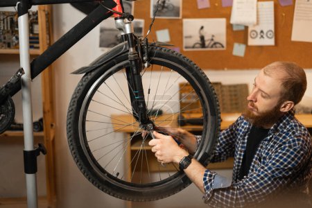 Bearded red-haired man in a checkered shirt repairs a bicycle while squatting in a garage or workshop. Side view