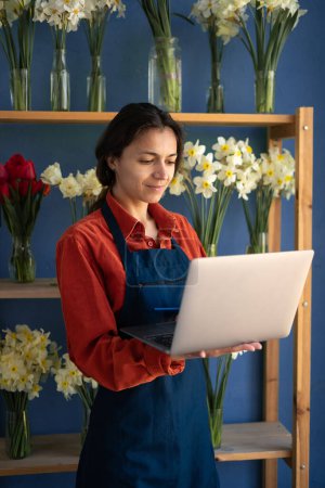 Florista hispana usando laptop en floristería. Trabajadora, propietaria de una pequeña empresa. Florista trabajando en la computadora de pie en su lugar de trabajo. Copiar espacio