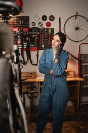 woman mechanic talking on phone, asking for help on repairing damaged bike, standing in garage or workshop. telephone call with bicycle engineer. Copy space