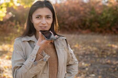 Hispanic millennial woman dictating audio message on smartphone walking in autumn park. Copy space